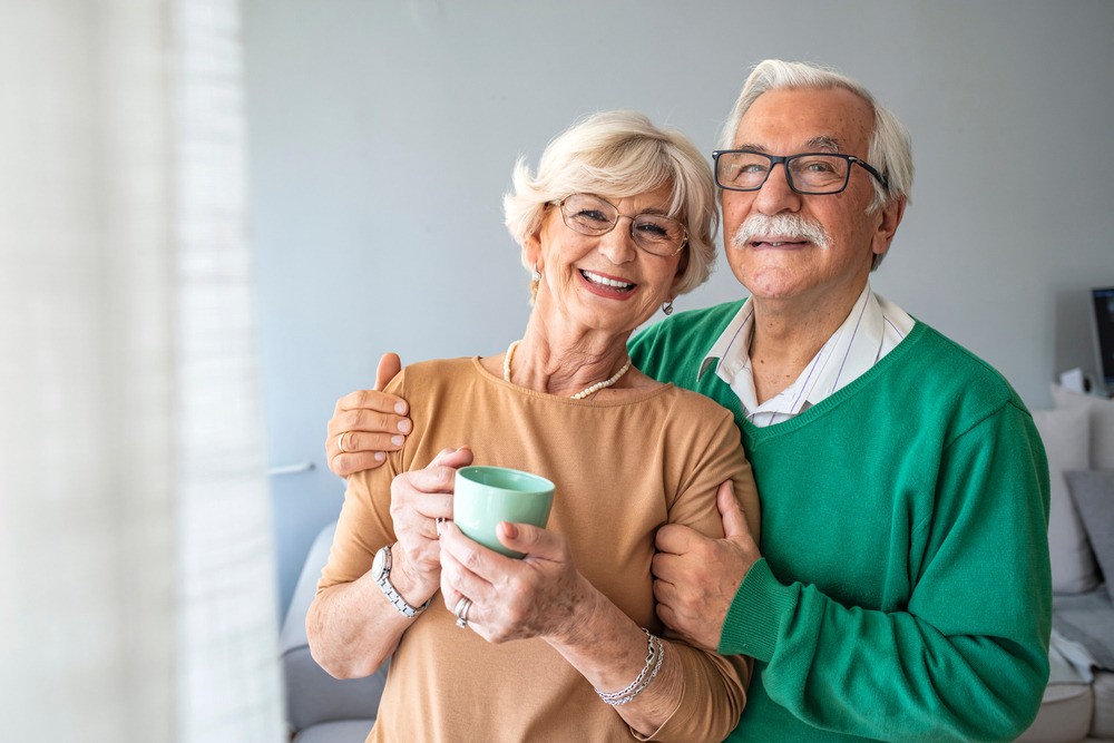 Happy,senior,man,embracing,woman,in,front,of,windows,at